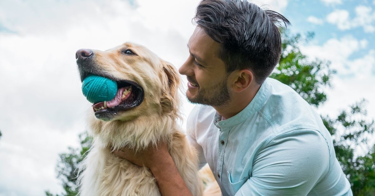 A man smiles as he pets a golden retriever holding a teal ball in its mouth, with a cloudy sky in the background.